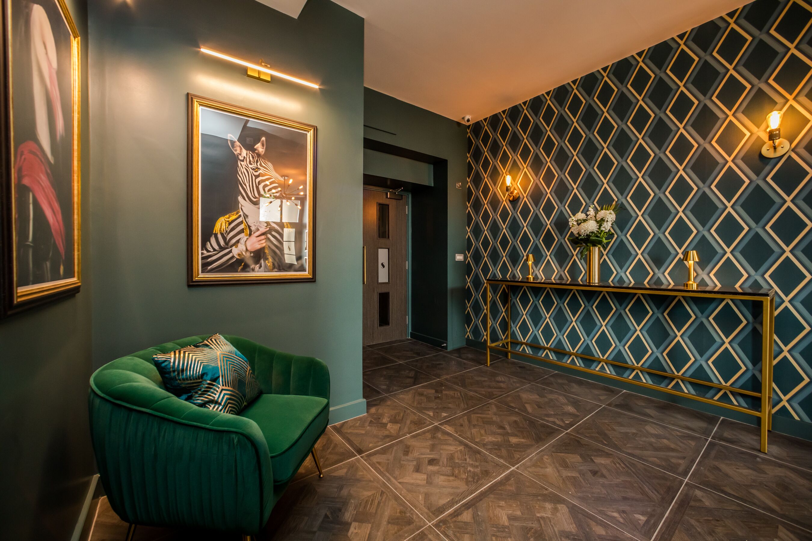 A modern interior with a green armchair, a framed zebra painting, and a patterned wall with a gold console.
