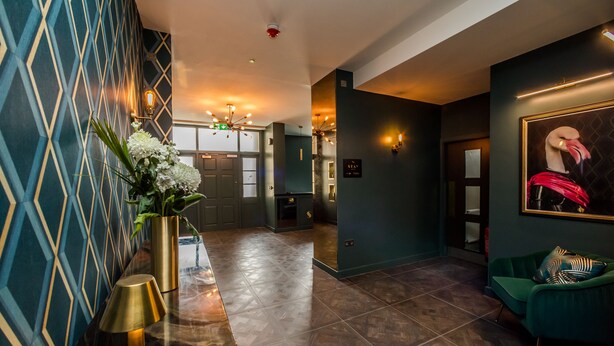 A modern lobby with a geometric patterned wall, a green sofa with patterned cushions, and a flamingo painting on the wall. - Stay Hotel (Huddersfield)