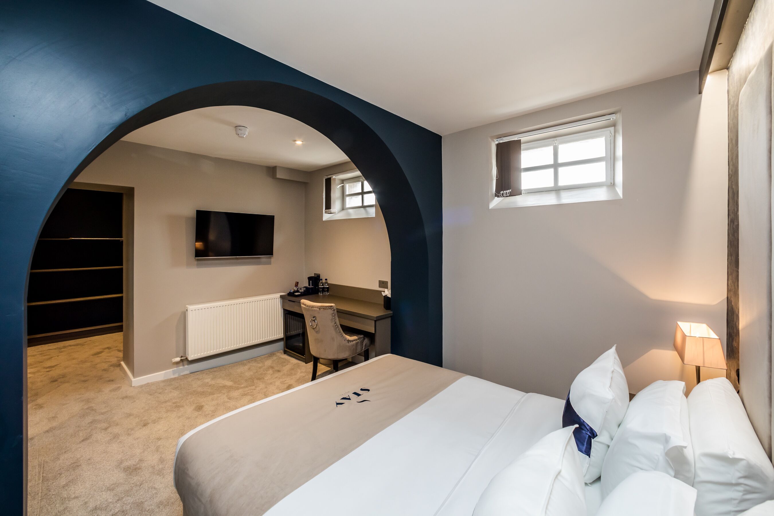 A modern hotel room with a large bed, a desk, a chair, a television, and a small window.