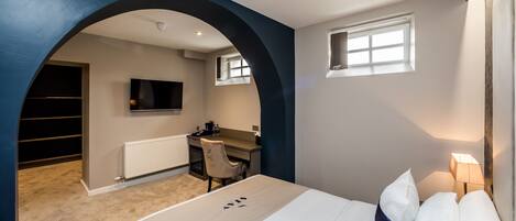 A modern hotel room with a large bed, a desk, a chair, a television, and a small window.