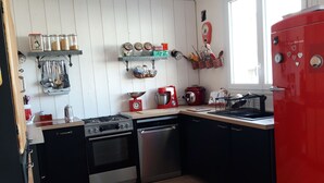 Una cocina con refrigerador rojo, electrodomésticos de acero inoxidable, piso de madera y paredes blancas con estantes que contienen utensilios de cocina y frascos.
