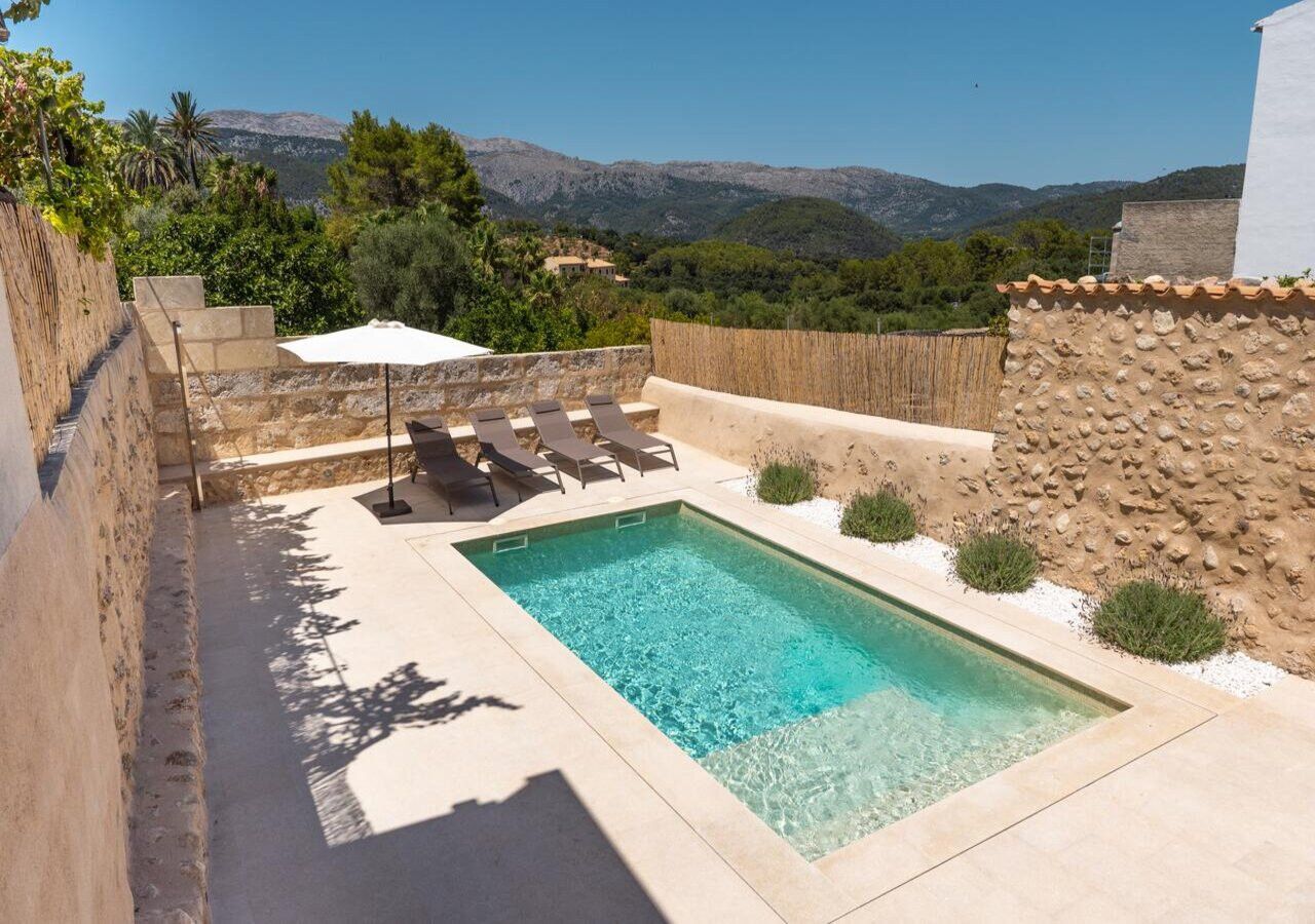 Ein rechteckiger Pool mit klarem Wasser, umgeben von einer Steinmauer und einem Holzzaun. In der Nähe des Pools stehen Liegestühle und ein weißer Sonnenschirm.