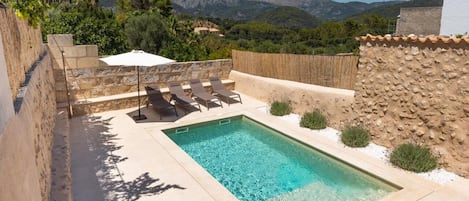 Ein rechteckiger Pool mit klarem Wasser, umgeben von einer Steinmauer und einem Holzzaun. In der Nähe des Pools stehen Liegestühle und ein weißer Sonnenschirm.