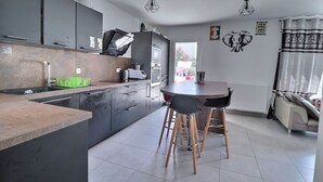 A modern kitchen with grey cabinets, a wooden countertop, and a dining area with two stools. - Villa en Location Près des Plages (Saint-Brès)