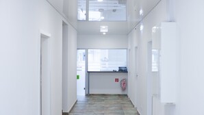 Een lichte, smalle gang met witte wanden en een betegelde vloer, die leidt naar een receptiegedeelte met een raam en een brandblusser.