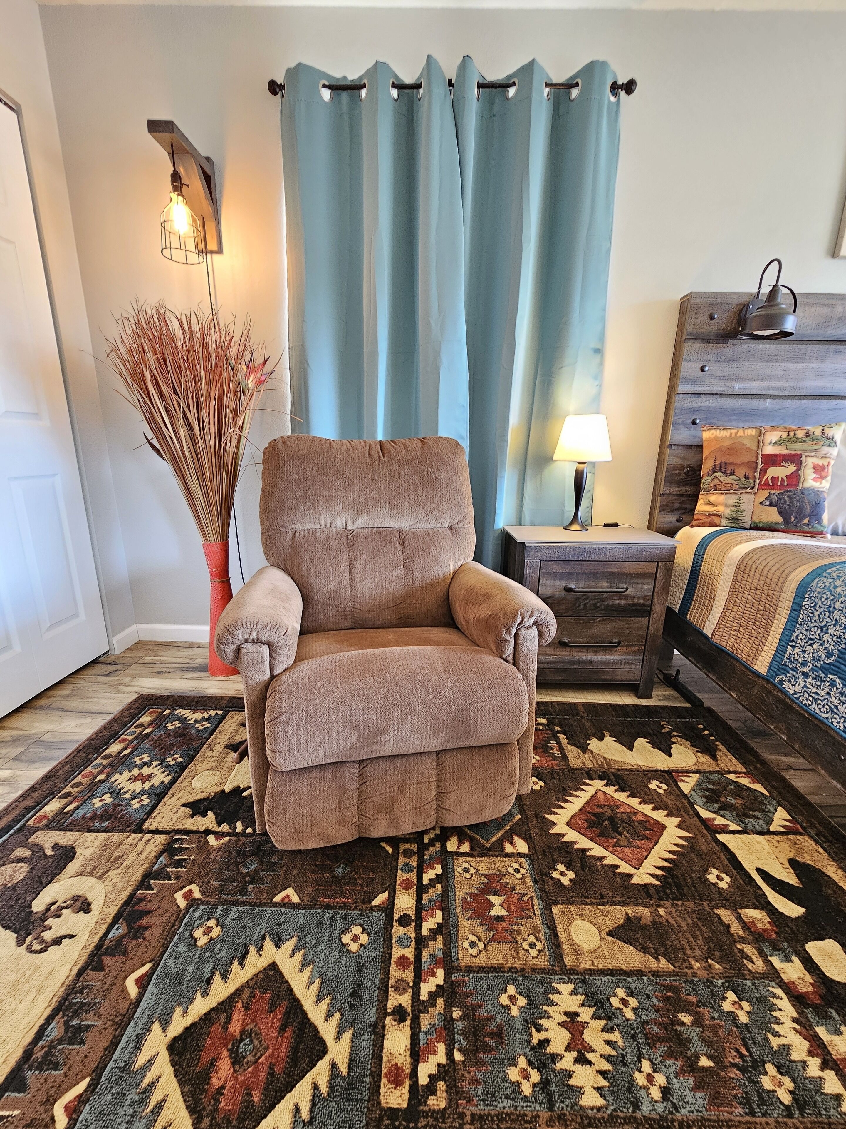 Una habitación con un sillón marrón, un tapete estampado, un aparador de madera, una cama con colcha azul y blanca, y una cortina azul.