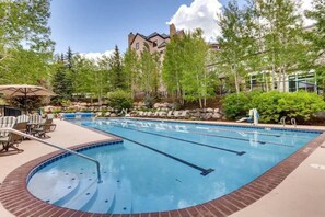 A swimming pool with multiple lanes, surrounded by lounge chairs and umbrellas, with a building in the background. - True Ski-In/Out - Highlands Slopeside Condo (Beaver Creek)