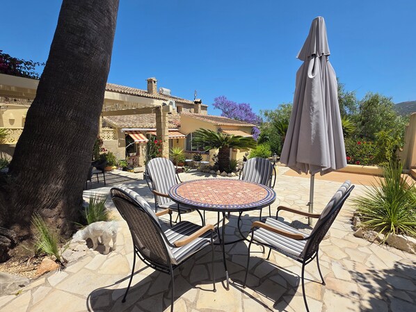An outdoor patio with a round table and chairs, surrounded by potted plants and a large umbrella.