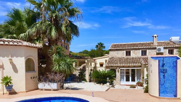 Una piscina con borde de azulejos, rodeada por un patio con plantas en macetas y un edificio de piedra con techo de tejas.