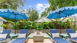 Un jardin avec quatre chaises longues bleues, un foyer central et deux parasols bleus qui offrent de l’ombre.