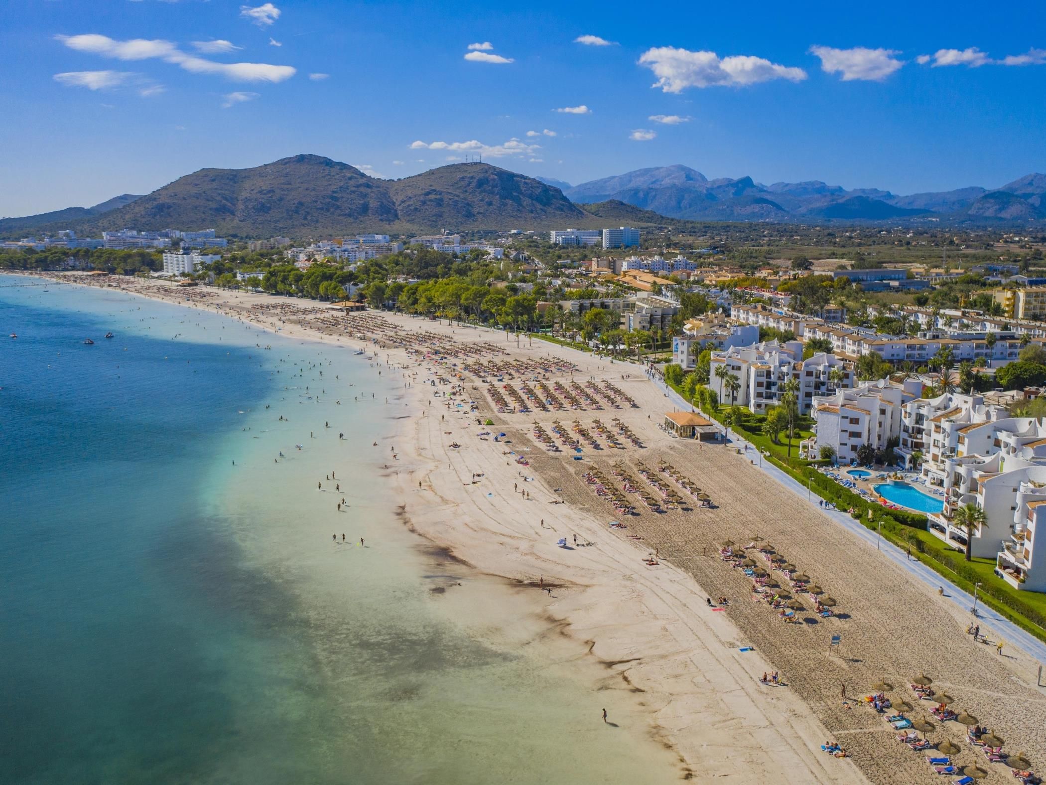 A coastal beach with a clear view of the sea, a sandy shore, and a row of buildings with balconies overlooking the beach.