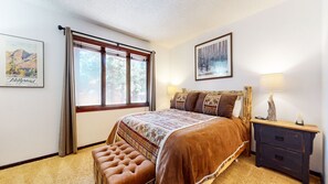 Quarto com cama grande, um banco, mesa de cabeceira, luminária e duas quadros nas paredes.