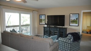 Een woonkamer met een bank, een geruit fauteuil, een televisie en een kast. Twee ingelijste foto's hangen aan de muur.