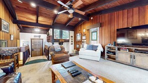 A cozy cabin interior with a wooden ceiling, a fireplace, a white sofa, a brown armchair, and a television.