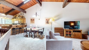 A spacious living area with a dining table, chairs, a sofa, a television, and a kitchen area with wooden cabinets and a hanging light fixture.