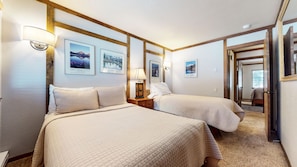 A hotel room with two beds, a nightstand with a lamp, framed pictures on the wall, and a door leading to another room.
