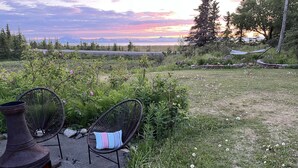 A serene outdoor seating area with two wicker chairs and a wood-burning stove, surrounded by lush greenery and a scenic view of mountains and a sunset. - Gorgeous Modern Alaskan Home with Ocean /Mt View 10 minute walk to sandy beach (Kenai)