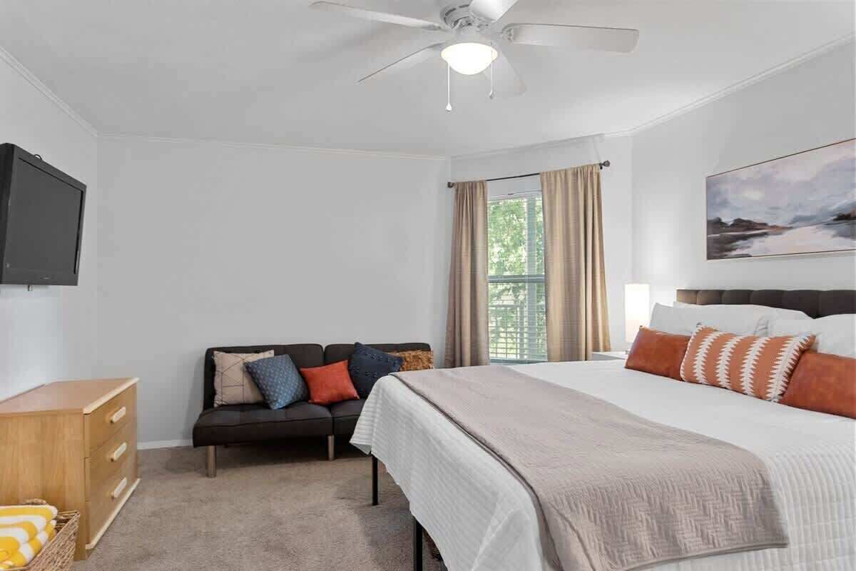 A bedroom with a bed, a sofa, a TV, a ceiling fan, and a window with curtains.