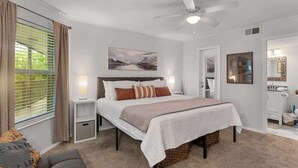 Una camera da letto con un letto, comodini, una ventola a soffitto, una finestra con tende, un quadro appeso al muro e un bagno con specchio e lavandino.