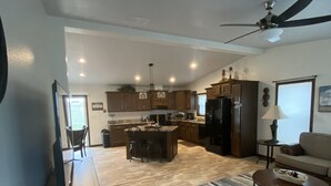 Amplia sala de estar con televisor grande, sofá y mesa de centro. Se observa una cocina con armarios de madera y mesa de comedor.