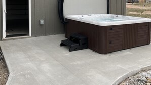 Un espacio tipo spa con un jacuzzi, una terraza de madera y un edificio con un ventanal grande y una puerta.