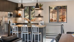 A modern kitchen with stainless steel appliances, a central island with bar stools, pendant lighting, and large windows offering a view of the outdoors.