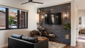 A modern living room with a black leather sofa, a flat-screen TV mounted on a brick wall, and a ceiling fan.