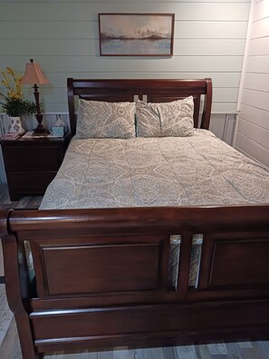 A wooden bed with a patterned bedspread, a nightstand with a lamp and decorative items, and a framed picture on the wall.