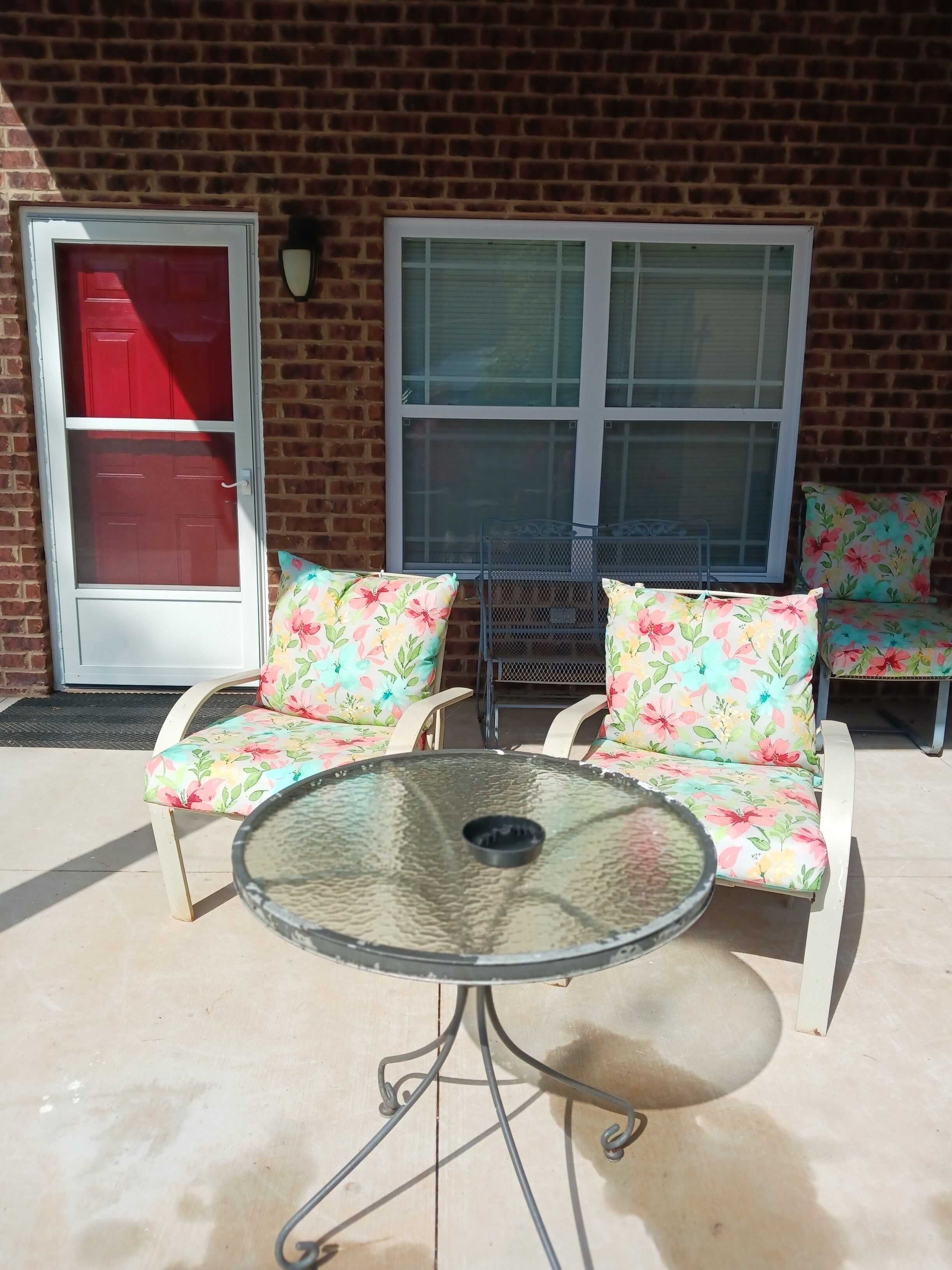 Un patio al aire libre con una mesa de vidrio y sillas con estampado floral, una puerta roja y un ventanal con persianas.