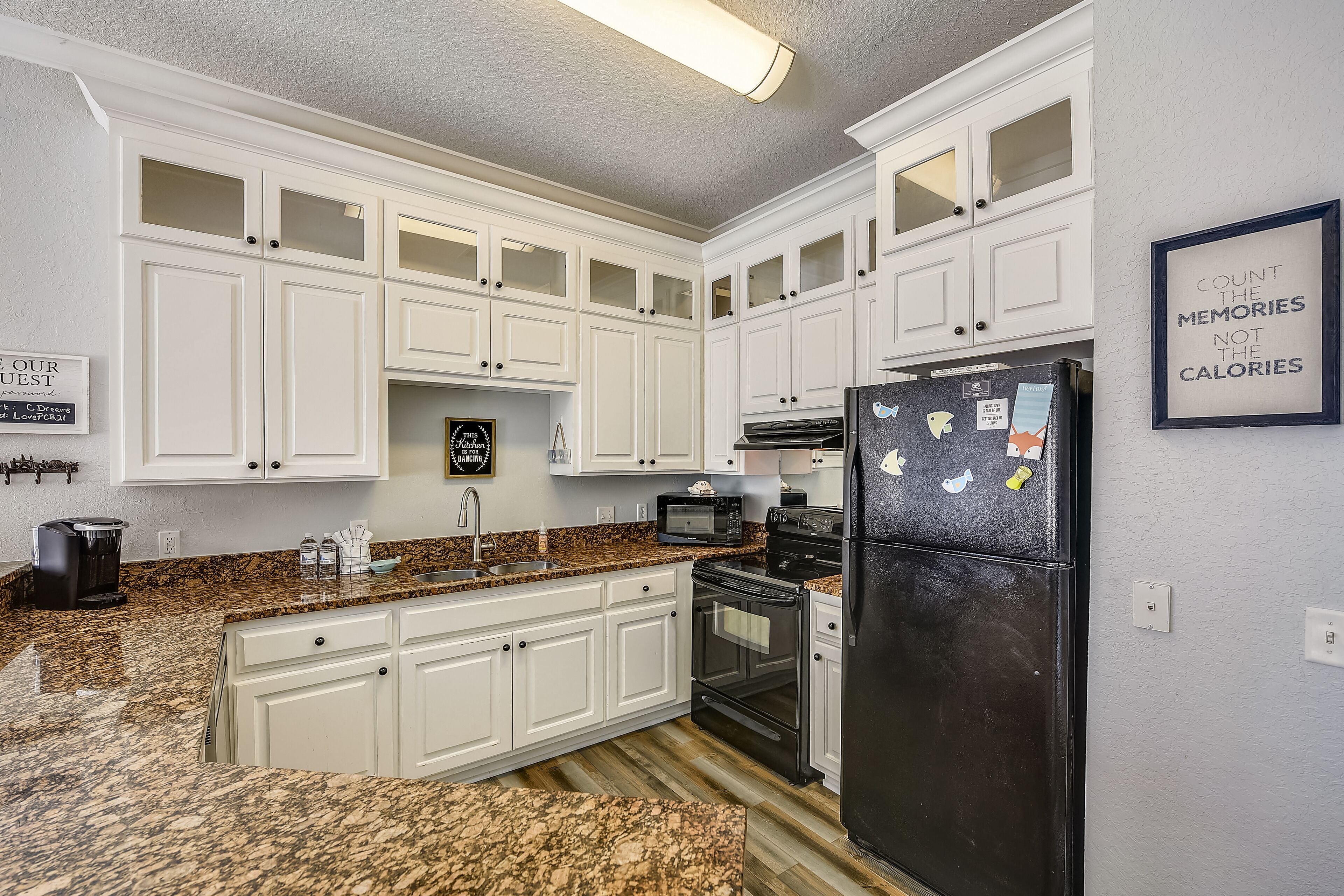 A kitchen with white cabinets, a black refrigerator, granite countertops, a microwave, a coffee maker, and a framed quote on the wall.