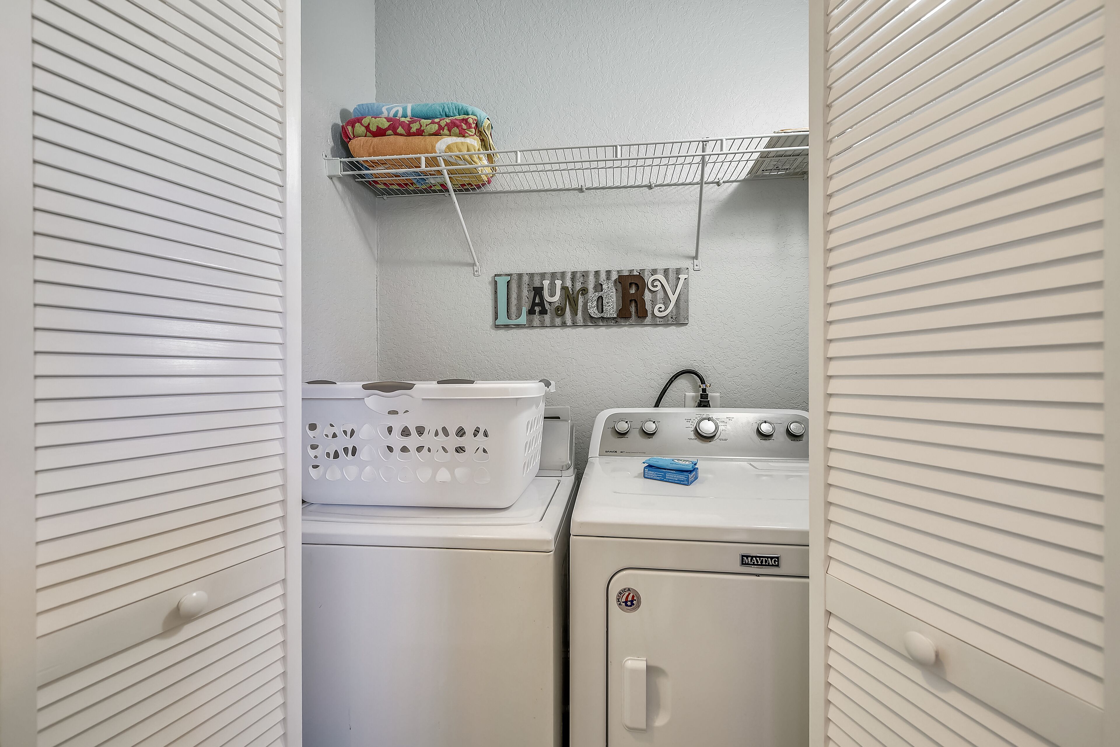 A laundry room with a white wall, a shelf with laundry supplies, and two stacked washing machines.