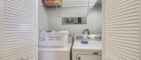 A laundry room with a white wall, a shelf with laundry supplies, and two stacked washing machines.