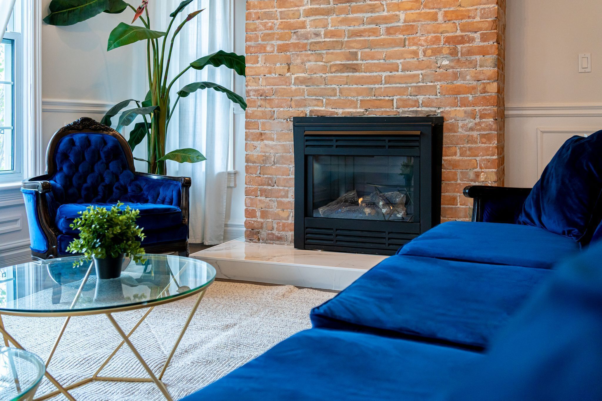 A living room with a brick fireplace, a blue sofa, a blue armchair, and a glass coffee table.