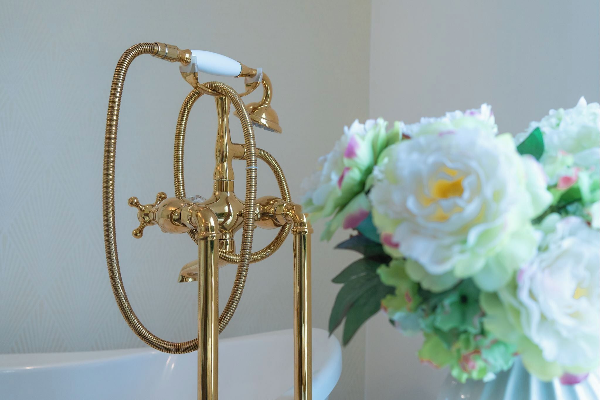 A bathroom with a gold showerhead and hose, a white bathtub, and a vase of flowers.