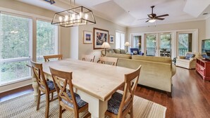 Un vaste espace de vie comprenant une table à manger, des chaises et un canapé. On y trouve également un ventilateur de plafond, une grande fenêtre avec des stores et un panneau indiquant la direction de la plage.