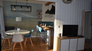 Eine moderne Küche mit Essbereich, Holzboden, einem runden Tisch, blauen Stühlen, einem Fernseher auf einem Holzgestell und einer Wand mit blauer und weißer Küchenrückwand.
