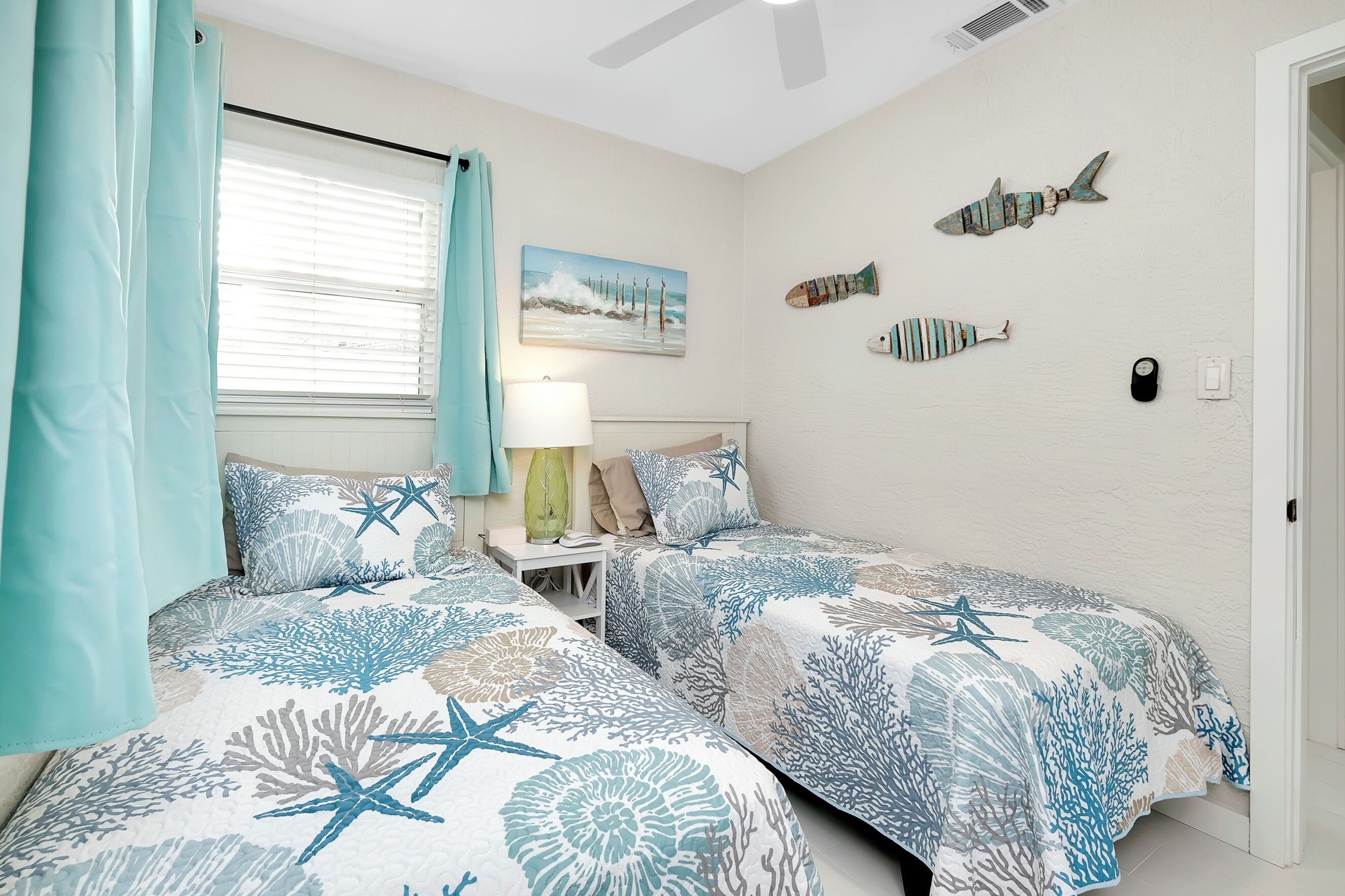 A bedroom with two beds, a window with curtains, a nightstand, a lamp, and wall art of fish and corals.