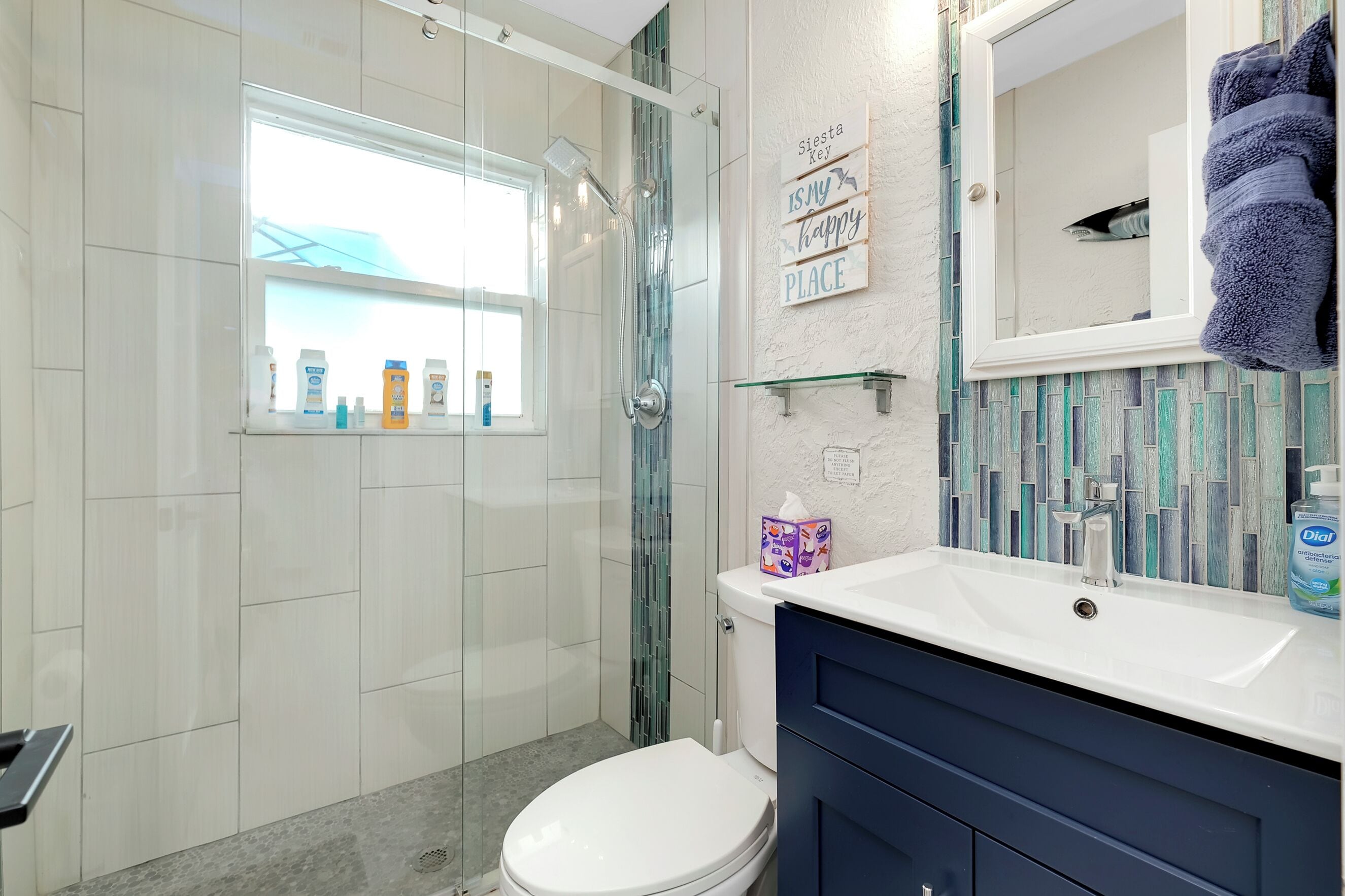 Un baño moderno con ducha de vidrio, inodoro blanco, lavamanos con espejo y un letrero decorativo en la pared.
