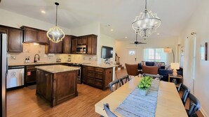 A spacious kitchen with dark wood cabinets, granite countertops, and a central island. There is a dining area with a long table and chairs, and a living room with a sofa and a TV.