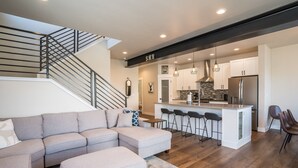 A modern living room with a sectional sofa, a kitchen island with bar stools, and a staircase with a metal railing. - New Family-Friendly Home Overlooking Fox Creek, Large Deck & Cozy Furnishings (Steamboat Springs)