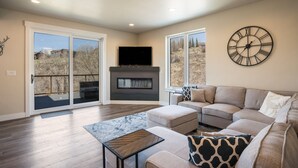 A spacious living room with a sectional sofa, a fireplace, a large wall clock, and sliding glass doors leading to a balcony. - New Family-Friendly Home Overlooking Fox Creek, Large Deck & Cozy Furnishings (Steamboat Springs)