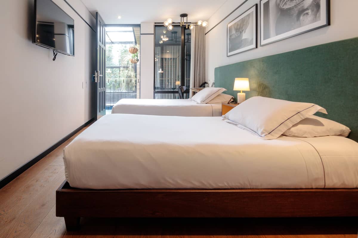 A hotel room with a bed, a TV, a desk, and a balcony with plants.