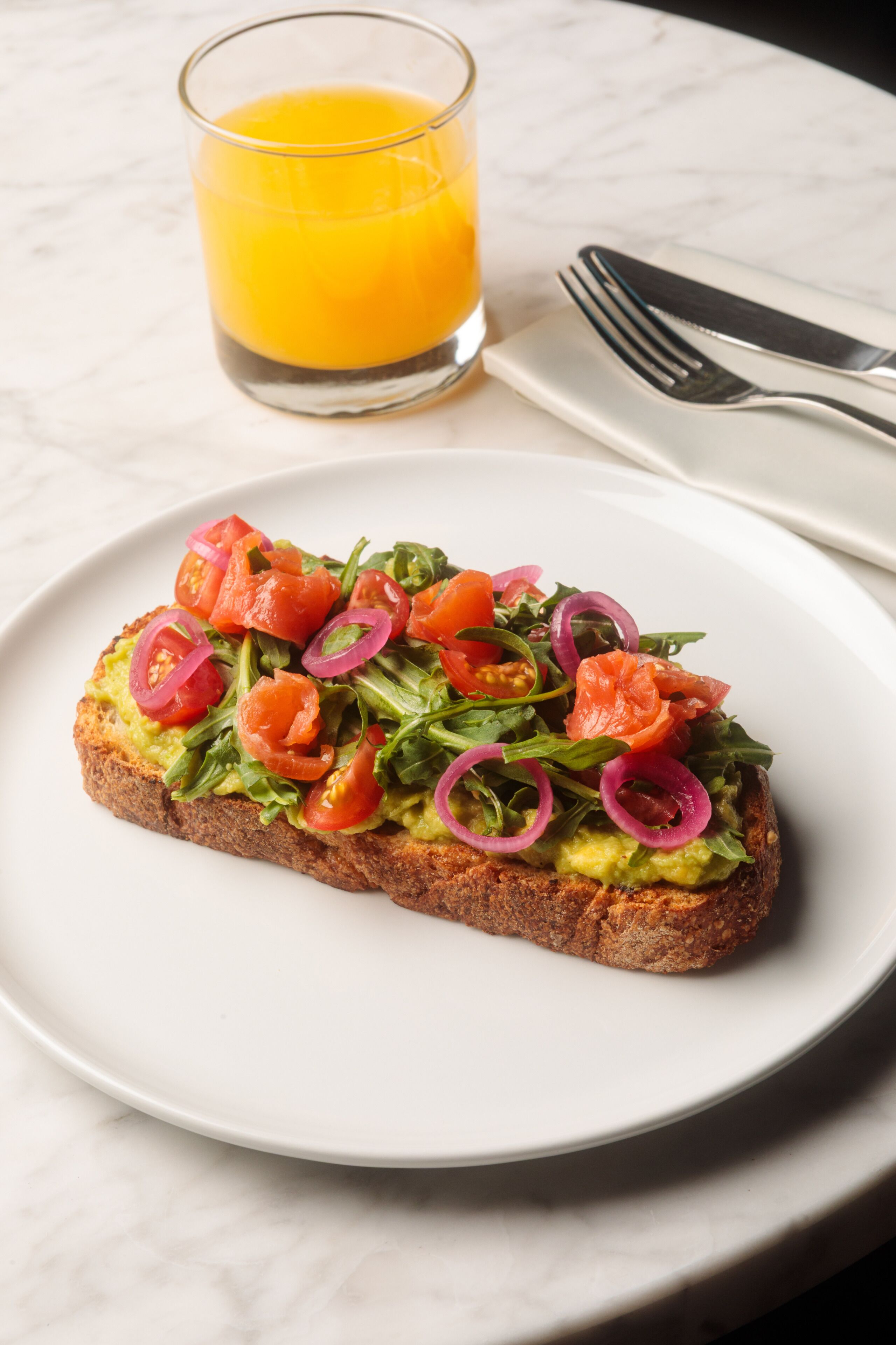 A plate with avocado toast topped with tomatoes and red onions, a glass of orange juice, and a fork and knife on a white plate.