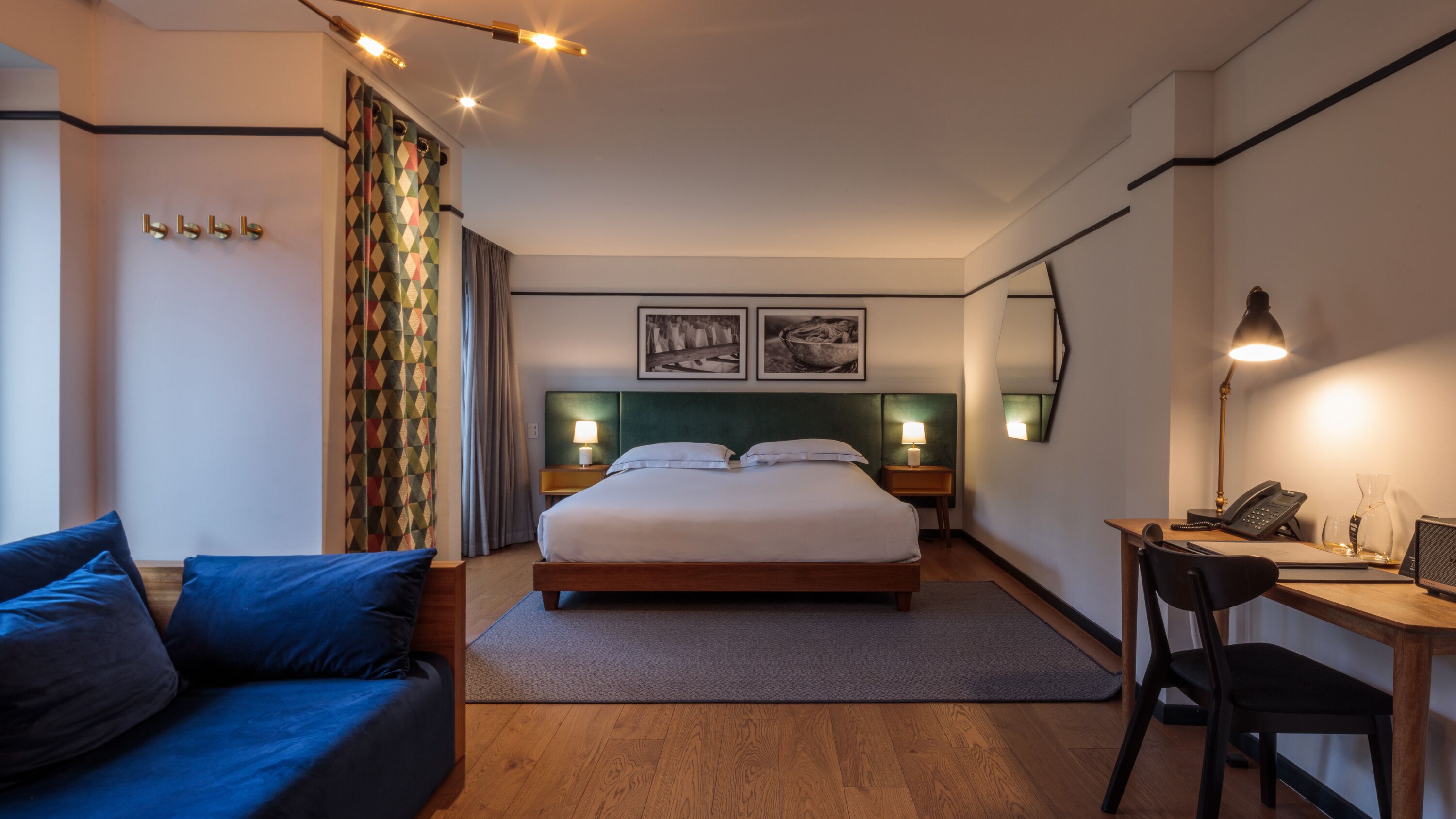 A hotel room with a bed, a desk with a phone, a chair, a couch, and a nightstand with lamps.
