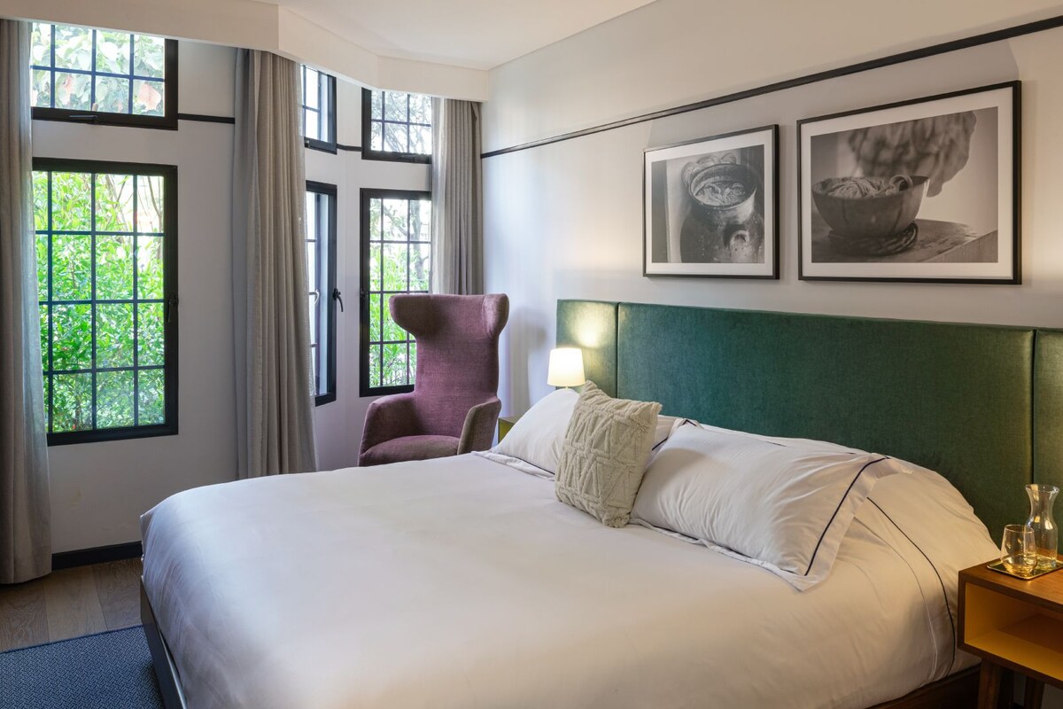 A hotel room with a large bed, a chair, two framed pictures, and a nightstand with a lamp.