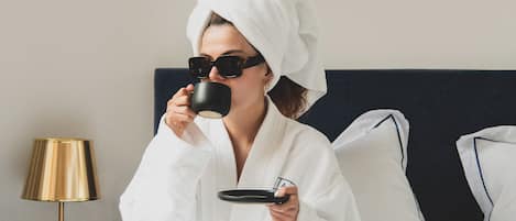 A person in a white bathrobe sitting on a bed with a tray of food, a lamp, and a framed picture on the wall.