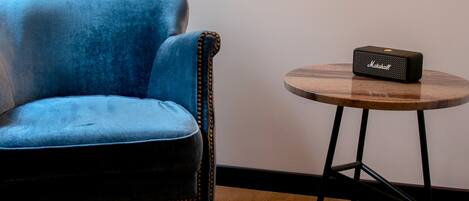 A room with a blue armchair, a small round table with a speaker, and a large mirror reflecting a window and curtains.
