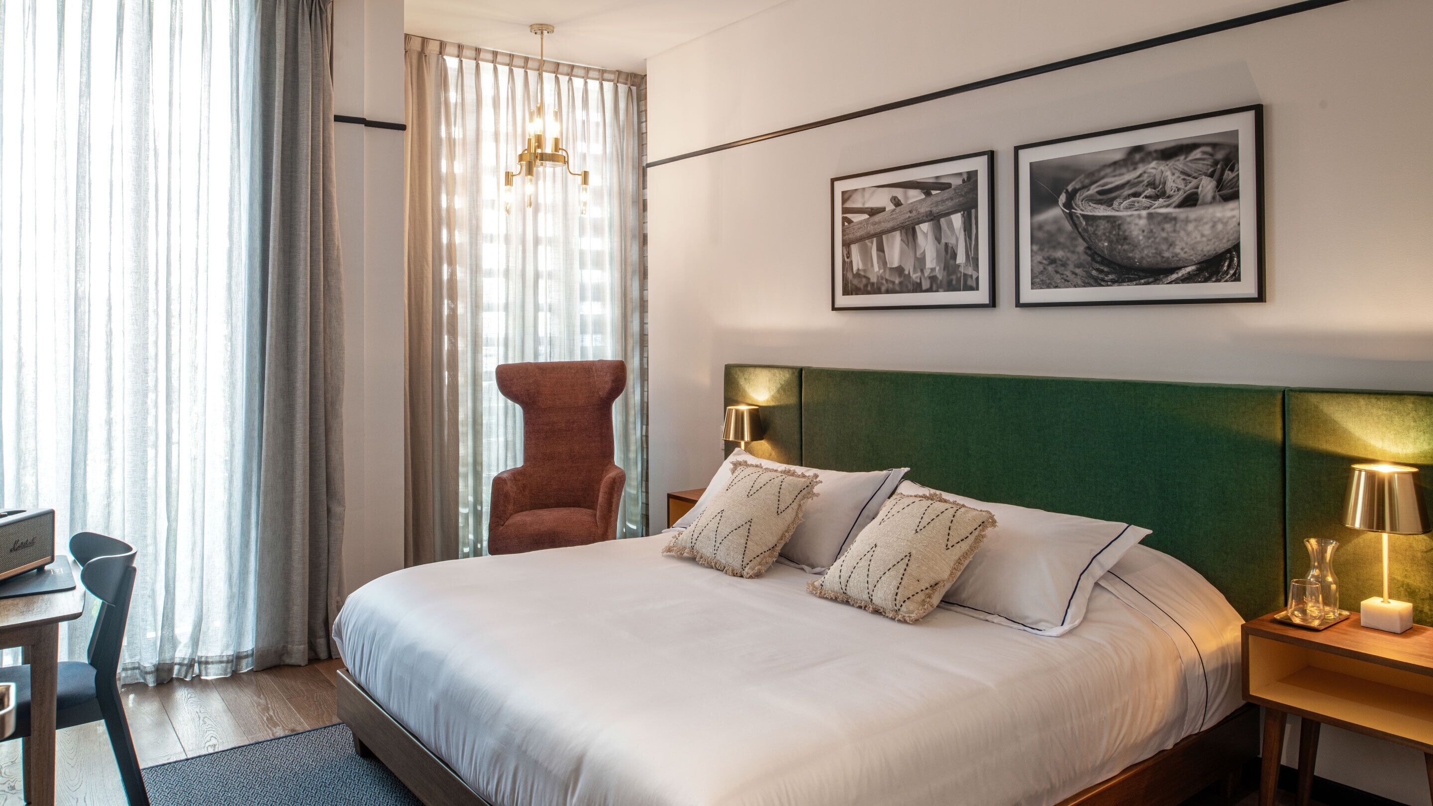 A hotel room with a large bed, a chair, a desk, and a window with curtains. There are two framed pictures on the wall.