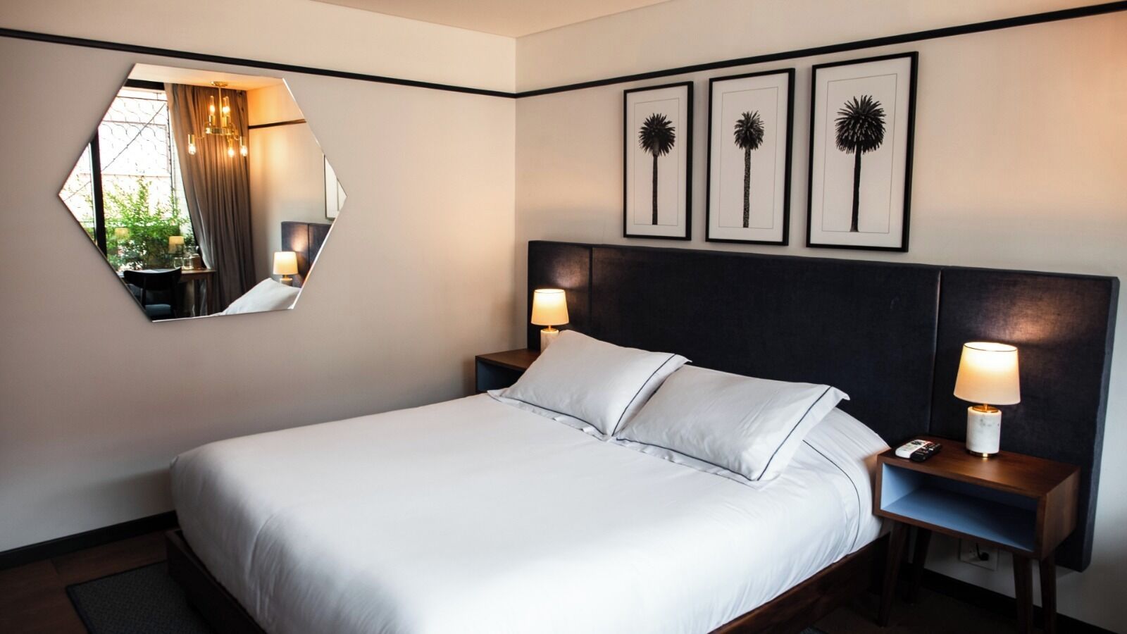 Quarto de hotel com uma cama, duas mesinhas de cabeceira com luminárias, um espelho redondo e três quadros na parede.