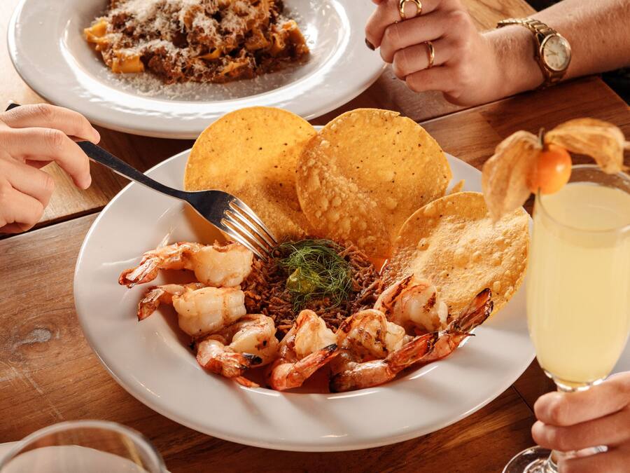 A dining table with plates of food, a glass of white wine, and a person wearing a watch.